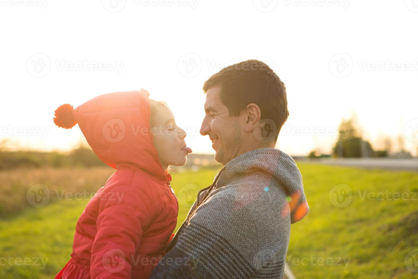 Little girl in red jacket with a hood hugs and stick out tongue her dad, smiles. Happy family, children's emotions, father's day, bright rays of the sun, Caucasian appearance. Space for text. photo