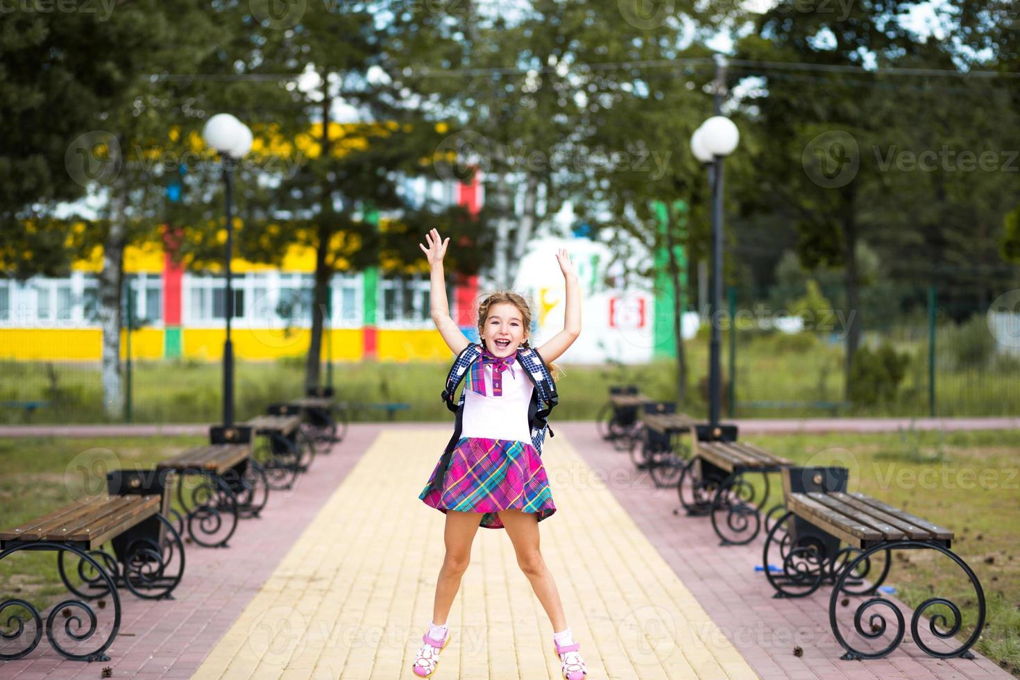 chica alegre con mochila y uniforme escolar en el patio de la escuela. regreso a la escuela, 1 de septiembre. alumno feliz salta de alegría y agita sus manos. educación primaria, estudiante de primaria foto