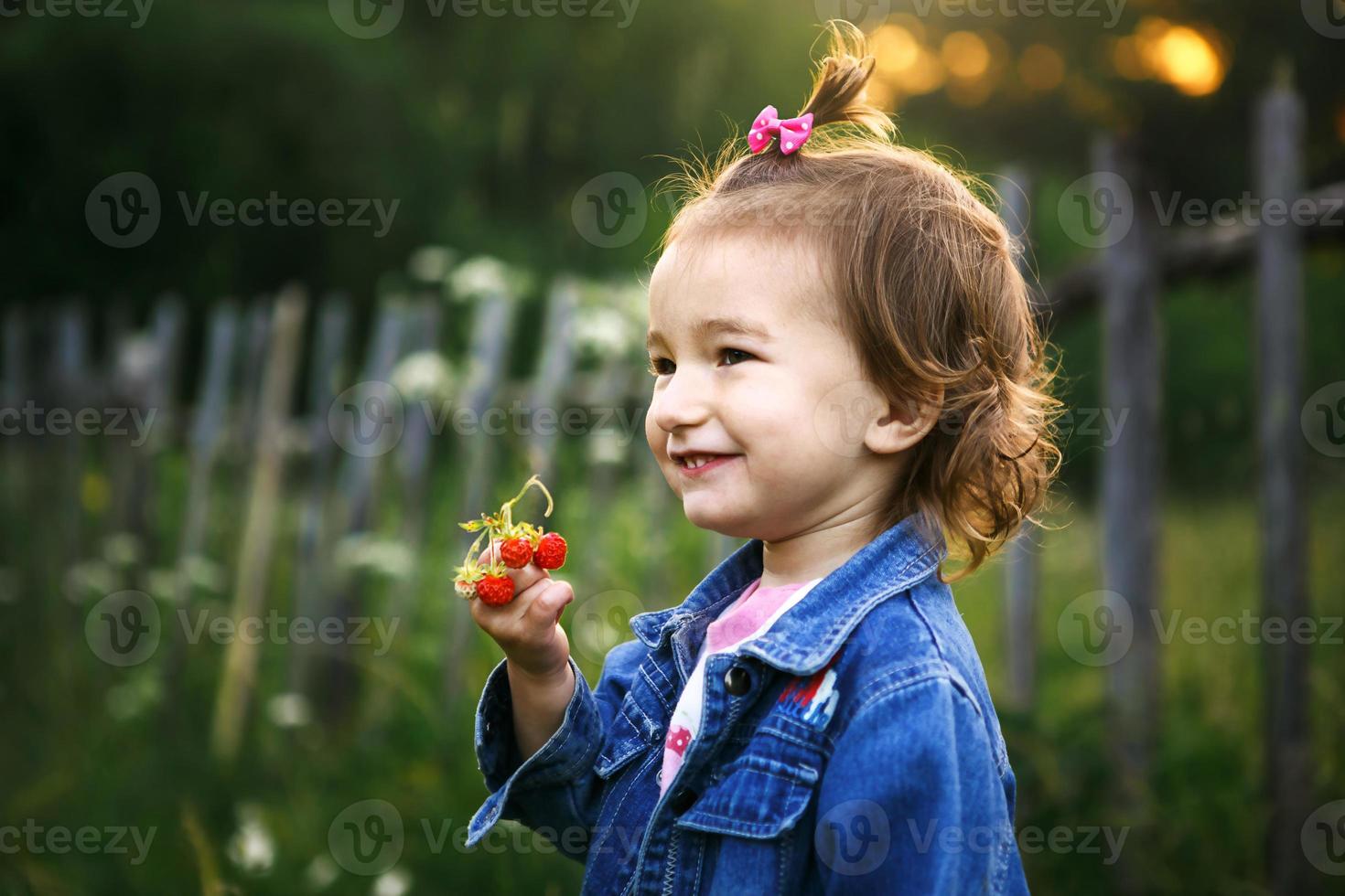 A little girl of 2 years old in the village holds a ripe strawberry in her hands and smiles slyly. Vitamins, summer time. International Children's Day. Nature, open air photo