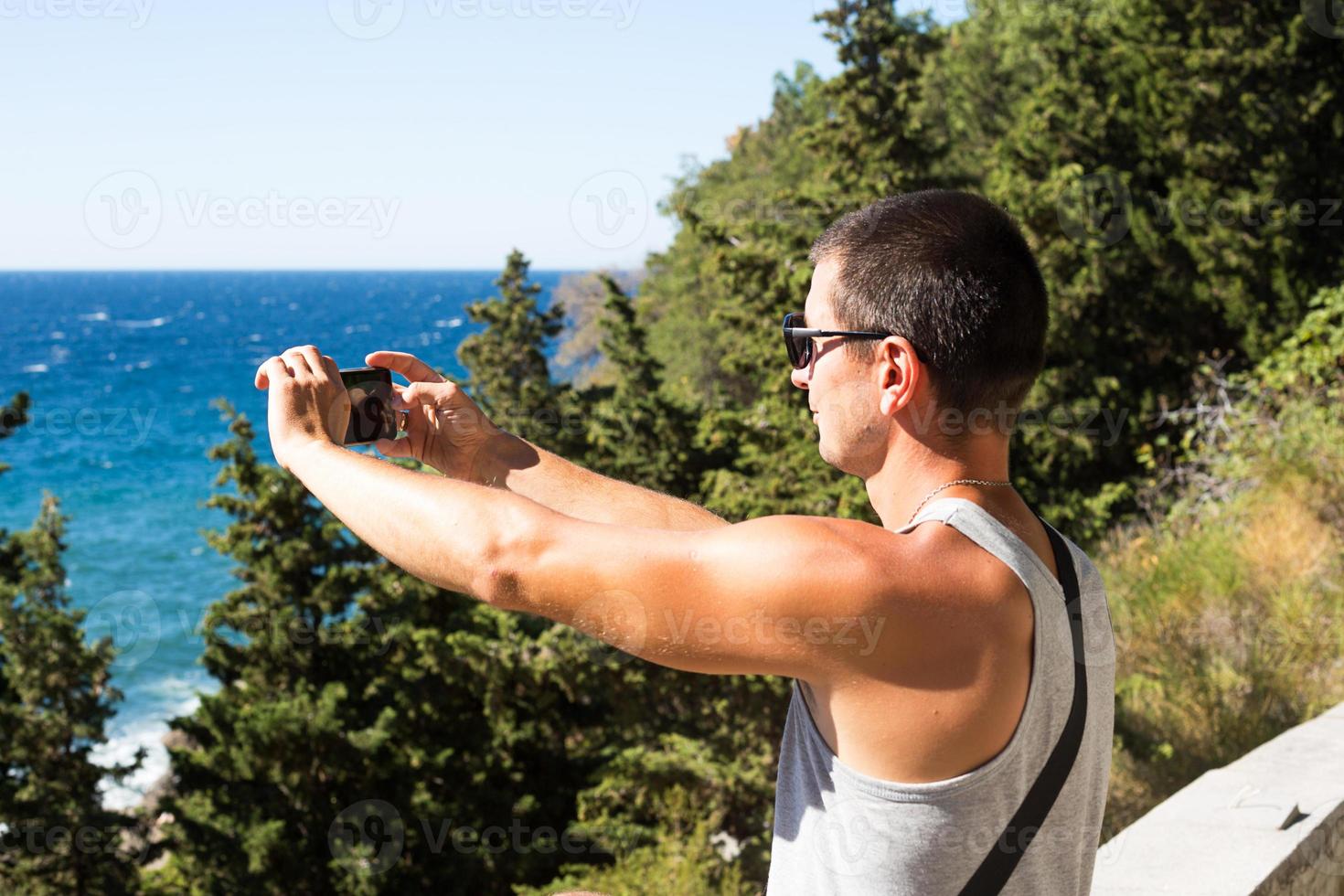 Male tourist takes photos on a smartphone of beautiful view of sea in a mountainous area from the top among coniferous trees. Video communication, using the phone when traveling. SIM card in roaming