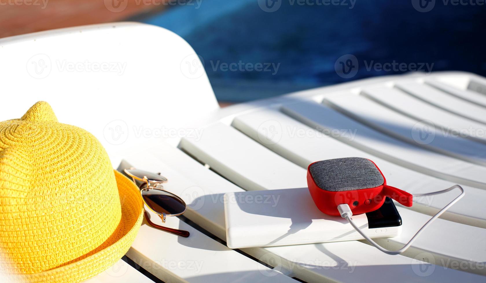 el altavoz portátil de música se carga desde el banco de energía a través de usb en una tumbona cerca de la piscina con accesorios de playa. siempre en contacto, dispositivos de viaje, batería externa para un teléfono inteligente. lugar para el texto. foto