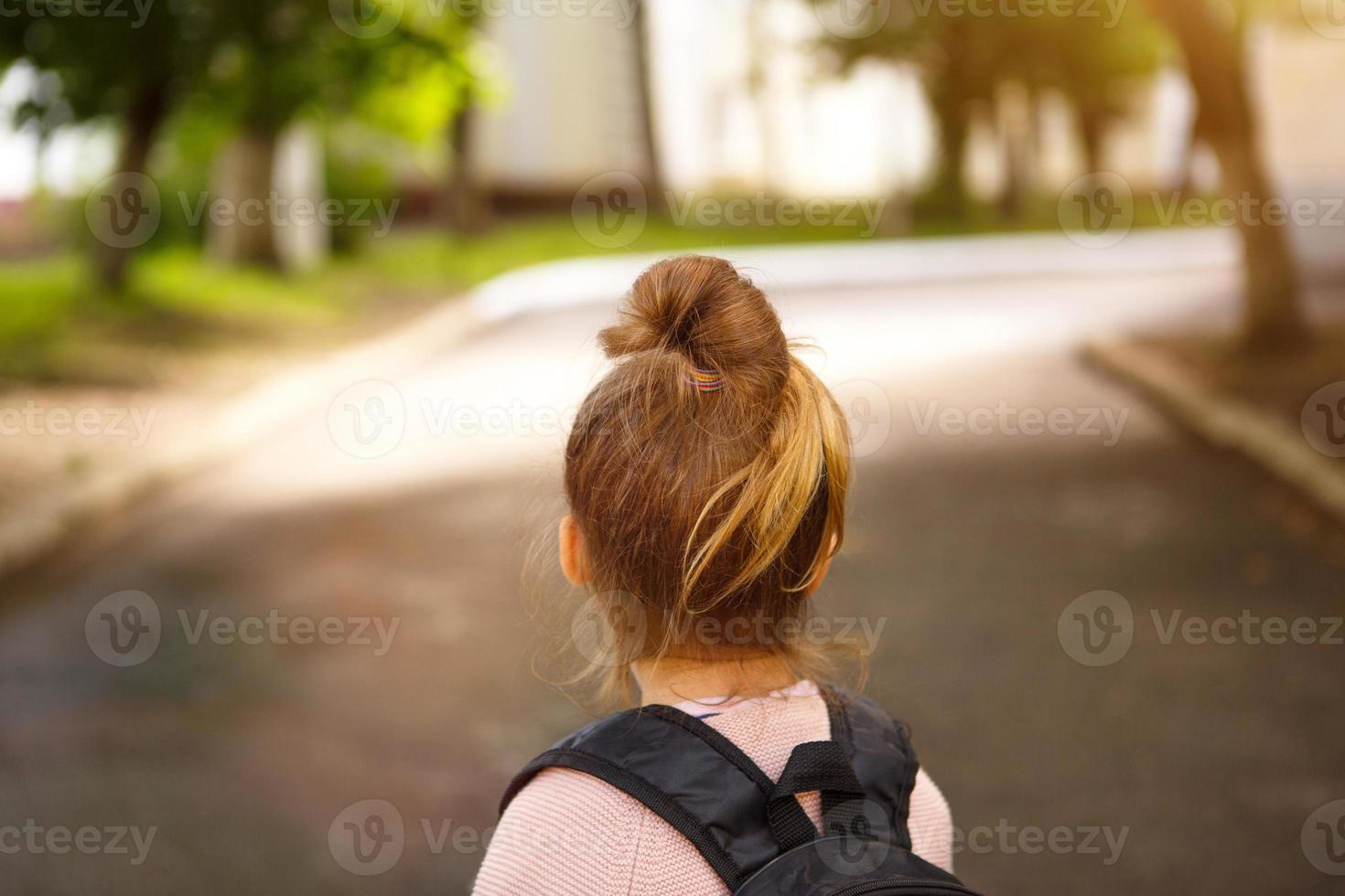 A little girl of Caucasian in a school uniform with a backpack looks at the road in the school yard. Concept back to school. Elementary school, developing activities for preschoolers. Space for text photo