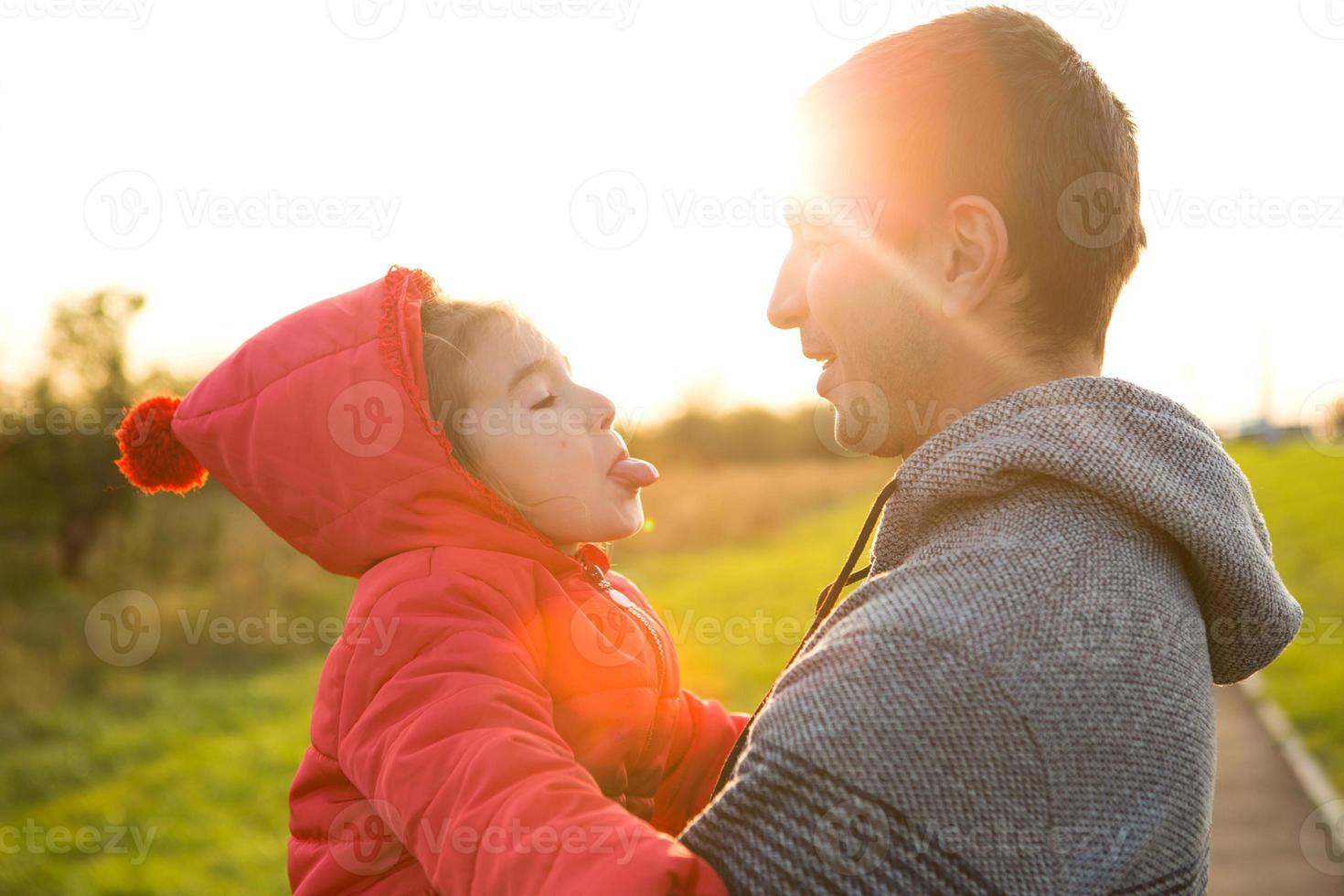Little girl in red jacket with a hood hugs and stick out tongue her dad, smiles. Happy family, children's emotions, father's day, bright rays of the sun, Caucasian appearance. Space for text. photo