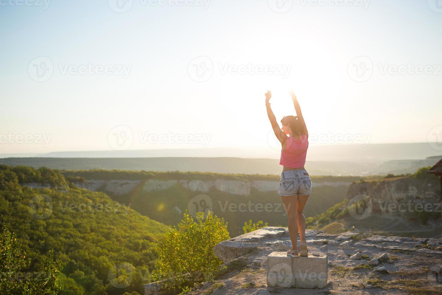 Female tourist with her hands raised looks at panoramic view on the top of the mountain and rejoices, enjoys it freedom and adventure. Trekking, travel, active ecotourism, healthy lifestyle, hiking photo