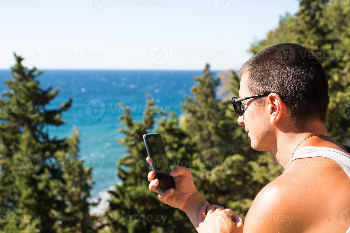 Male tourist takes photos on a smartphone of beautiful view of sea in a mountainous area from the top among coniferous trees. Video communication, using the phone when traveling. SIM card in roaming