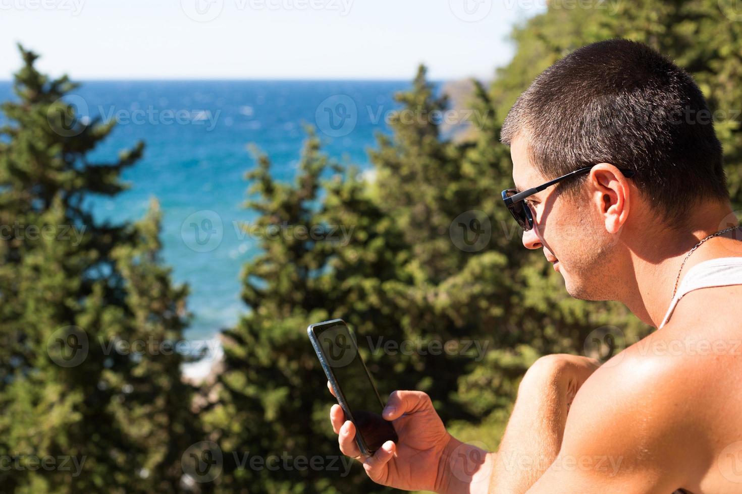 Male tourist takes photos on a smartphone of beautiful view of sea in a mountainous area from the top among coniferous trees. Video communication, using the phone when traveling. SIM card in roaming