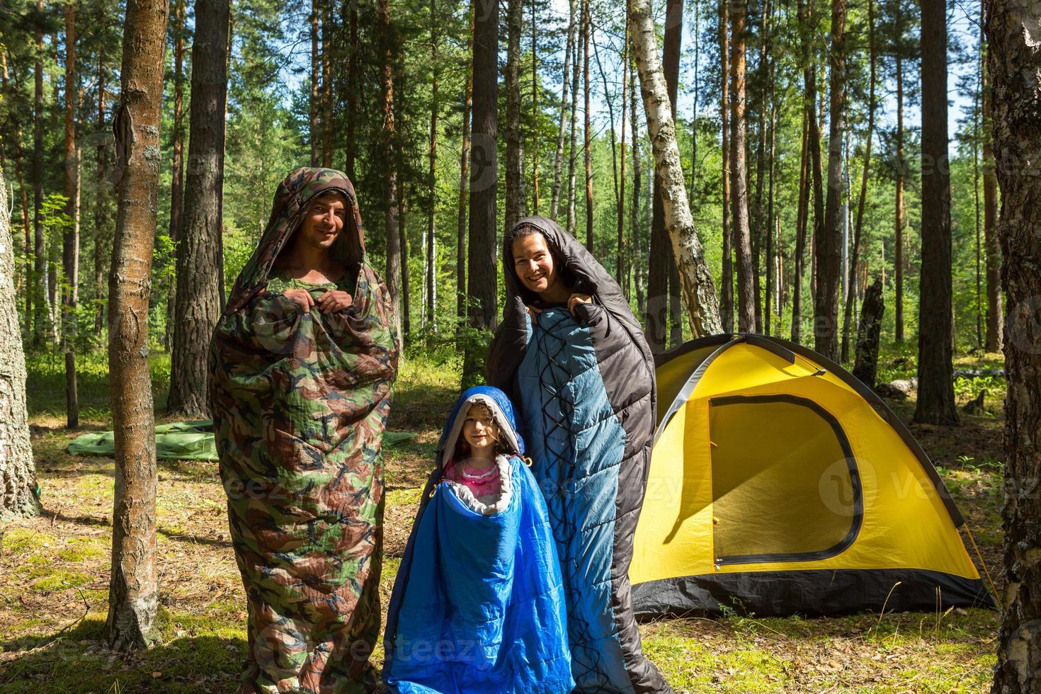 Family of tourists from a father, mother and little daughter pose in sleeping bags near a tent. Family outdoor recreation, domestic tourism, camping, hiking equipment. Pupated like caterpillars-humor photo