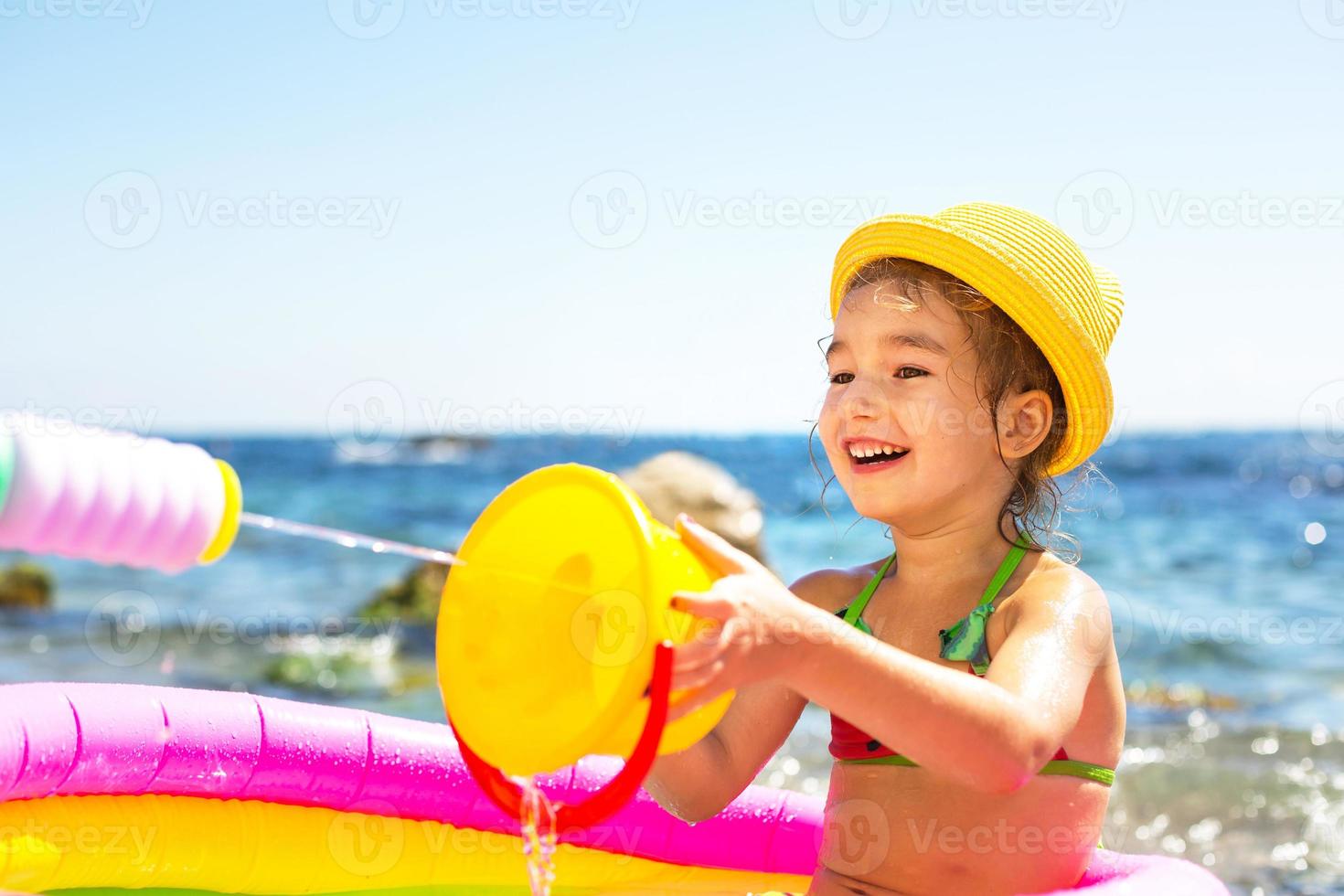 chica con sombrero de paja amarillo juega con el viento, el agua y un dispensador de agua en una piscina inflable en la playa. productos indelebles para proteger la piel de los niños del sol, las quemaduras solares. recurso en el mar. foto