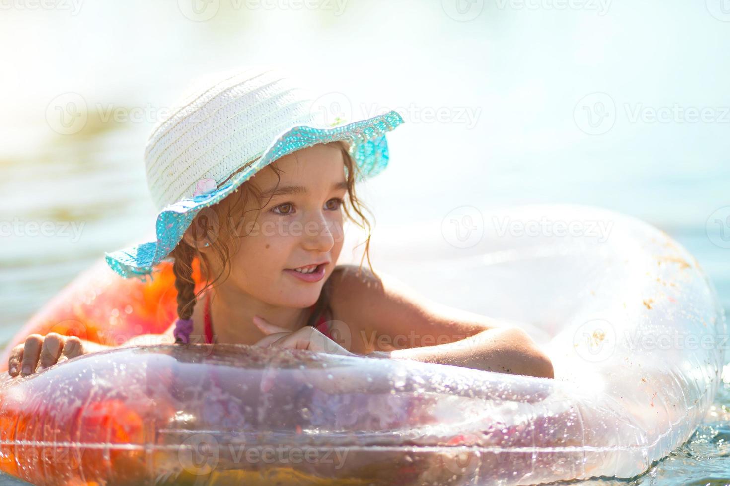 chica con sombrero nadando en el río con un círculo inflable transparente en forma de corazón con plumas naranjas dentro.el mar con un fondo arenoso. vacaciones en la playa, nadar, broncearse, protectores solares. foto