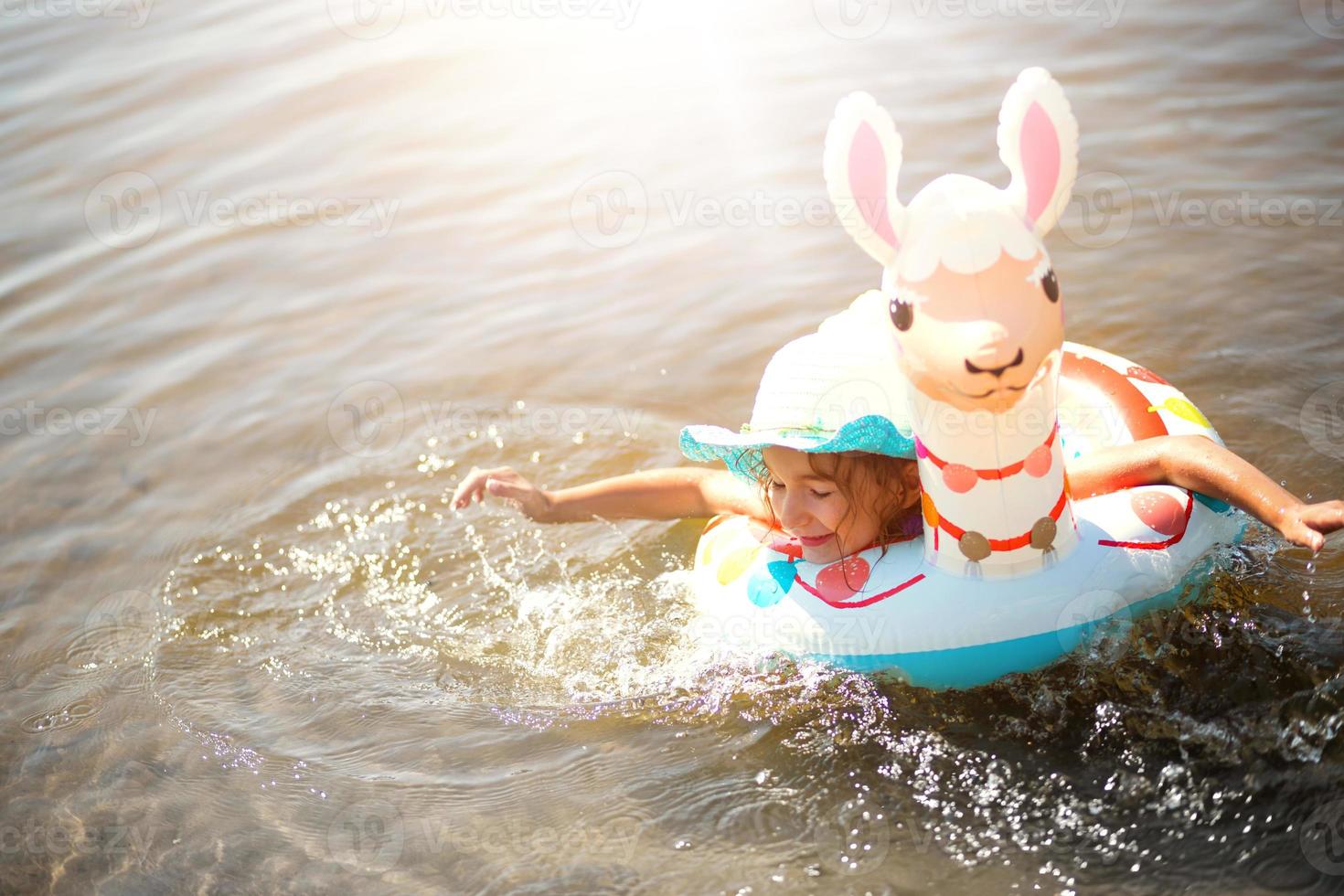 Girl in a hat swimming in the river with inflatable circle in the shape of a lama. Inflatable alpaca for a child. Sea with a sandy bottom. Beach holidays, swimming, tanning, sunscreens. photo