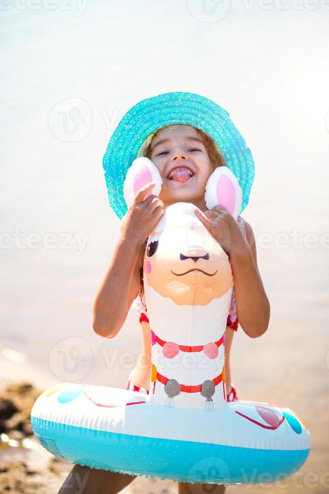 Girl in a hat it stands on the shore with inflatable circle in the shape of a lama. Inflatable alpaca for a child. Sea with a sandy bottom. Beach holidays, swimming, tanning, sunscreens. photo