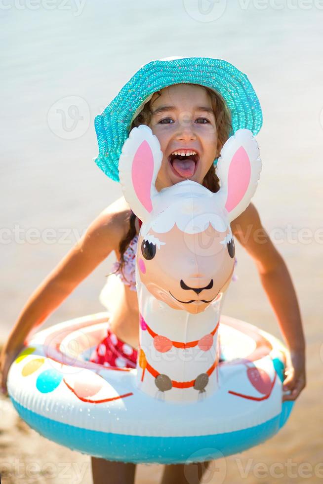 Girl in a hat it stands on the shore with inflatable circle in the shape of a lama. Inflatable alpaca for a child. Sea with a sandy bottom. Beach holidays, swimming, tanning, sunscreens. photo