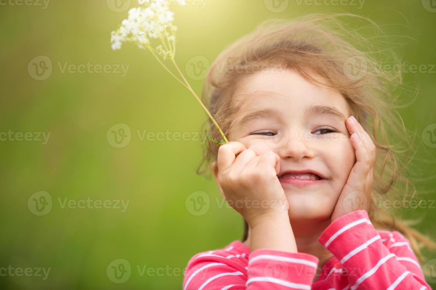 Little girl in a coral striped T-shirt on a green background in a field holds her face in her hands and smiles slyly. Children's Day, happy child, environmental and nature protection, insect repellent photo