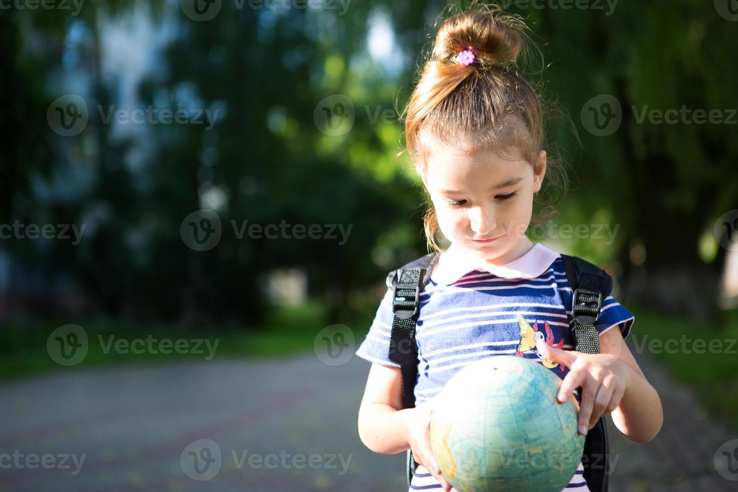 A girl from an elementary school of Caucasian appearance with a backpack on her shoulders holds a globe in her hands and carefully studies it. Education, return to school, September 1. Copy space photo