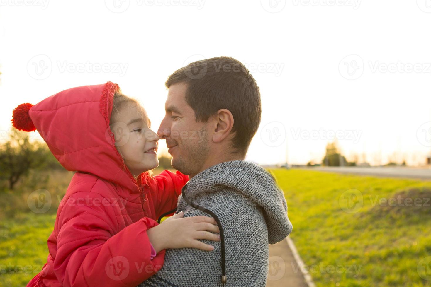 Little girl in red jacket with a hood hugs and kisses her dad, smiles, touches her nose. Happy family, children's emotions, father's day, bright rays of the sun, Caucasian appearance. Space for text. photo