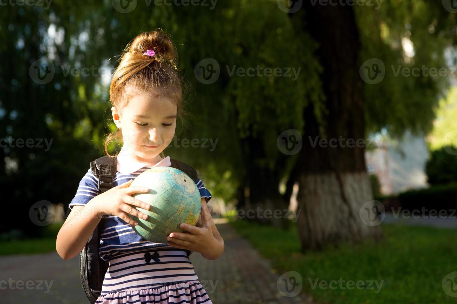 A girl from an elementary school of Caucasian appearance with a backpack on her shoulders holds a globe in her hands and carefully studies it. Education, return to school, September 1. Copy space photo