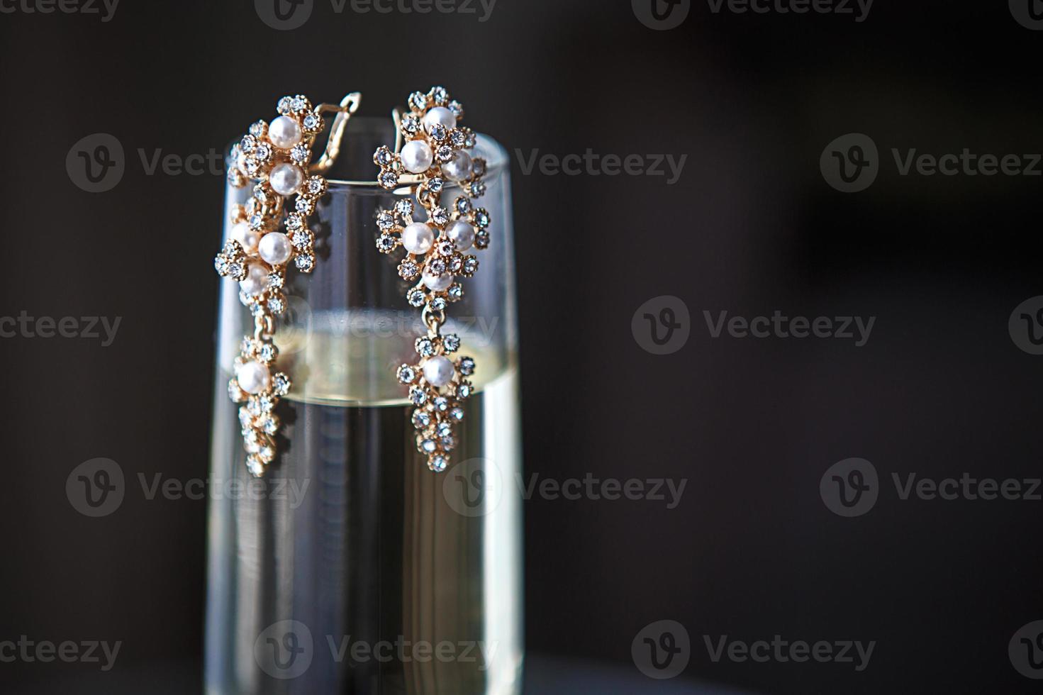 Large gold earrings with pearls and crystals on a clear glass of champagne. Morning and gathering of the bride, costume jewelry for the wedding celebration. Jewelry for the holiday. Black background, photo