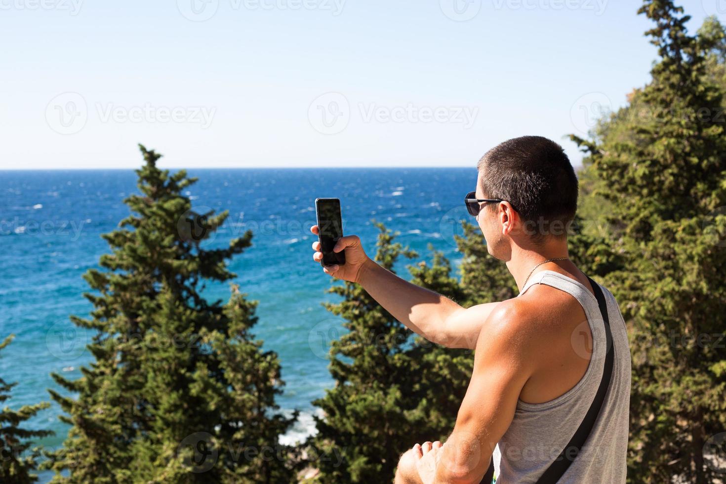 Male tourist takes photos on a smartphone of beautiful view of sea in a mountainous area from the top among coniferous trees. Video communication, using the phone when traveling. SIM card in roaming
