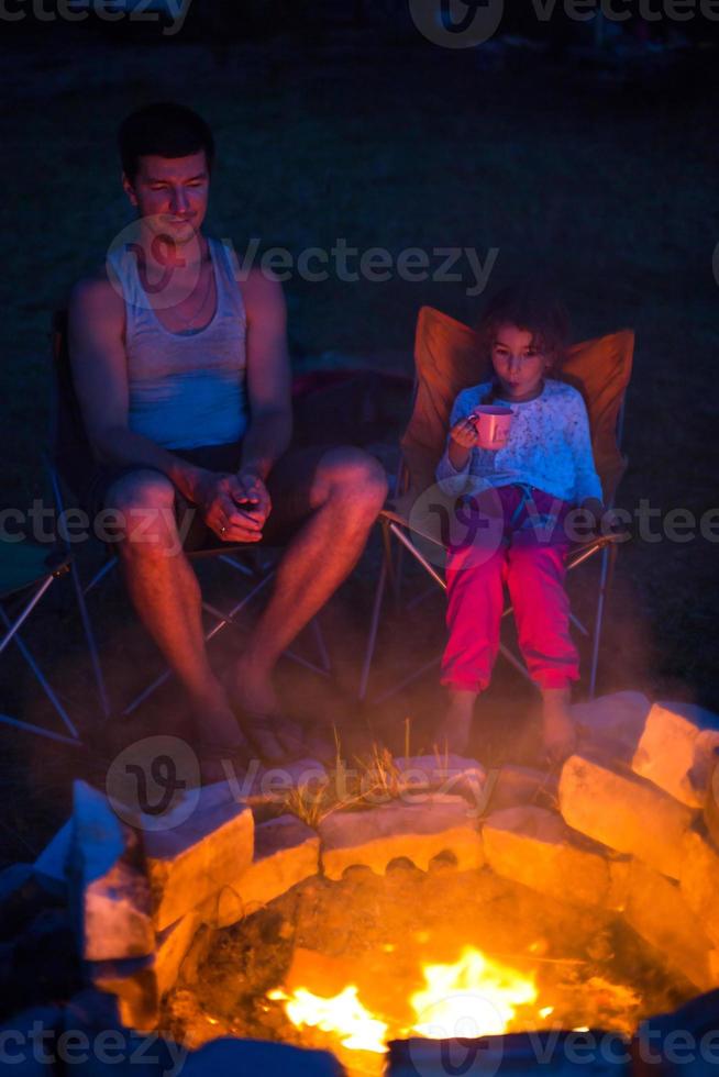 Dad and daughter sit at night by the fire in the open air in the summer in nature. Family camping trip, gatherings around the campfire. Father's Day, barbecue. Camping lantern and tent photo