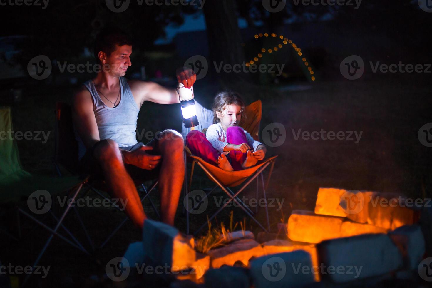 Dad and daughter sit at night by the fire in the open air in the summer in nature. Family camping trip, gatherings around the campfire. Father's Day, barbecue. Camping lantern and tent photo