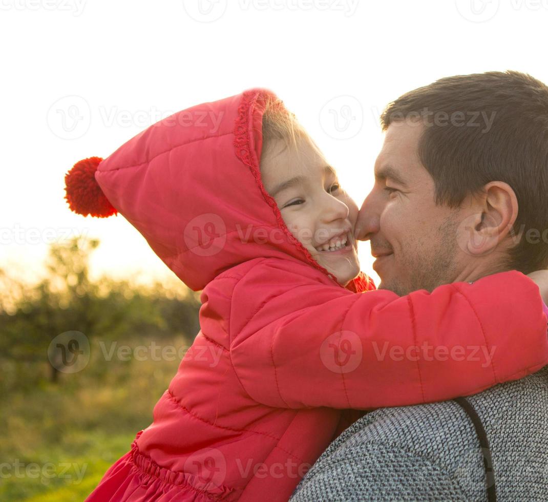 niñita con chaqueta roja con capucha abraza y besa a su papá, sonríe, toca su nariz. familia feliz, emociones de los niños, día del padre, rayos brillantes del sol, apariencia caucásica. espacio para texto. foto