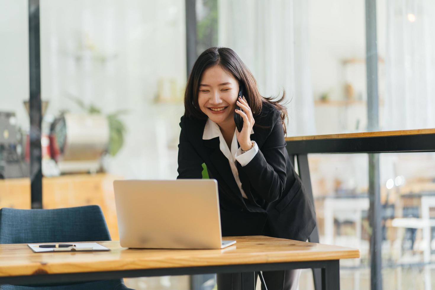 Happy smiling Asian business woman working on laptop at office, using smart phone. Businesswoman sitting at her working place photo