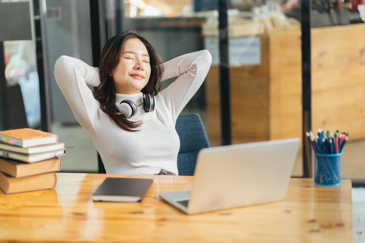 Happy calm asian girl student relaxing holding hands behind finished study work breathing fresh air sit at home office desk feel stress relief stretching doing exercise dreaming enjoy peace of mind photo