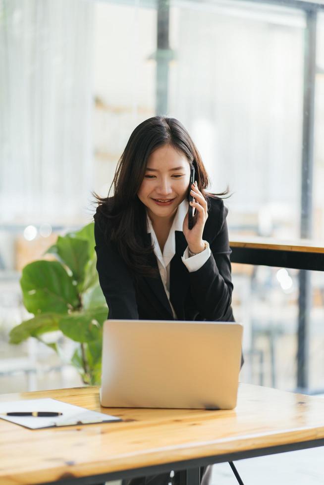 mujer de negocios asiática sonriente feliz trabajando en una computadora portátil en la oficina, usando un teléfono inteligente. mujer de negocios sentada en su lugar de trabajo foto