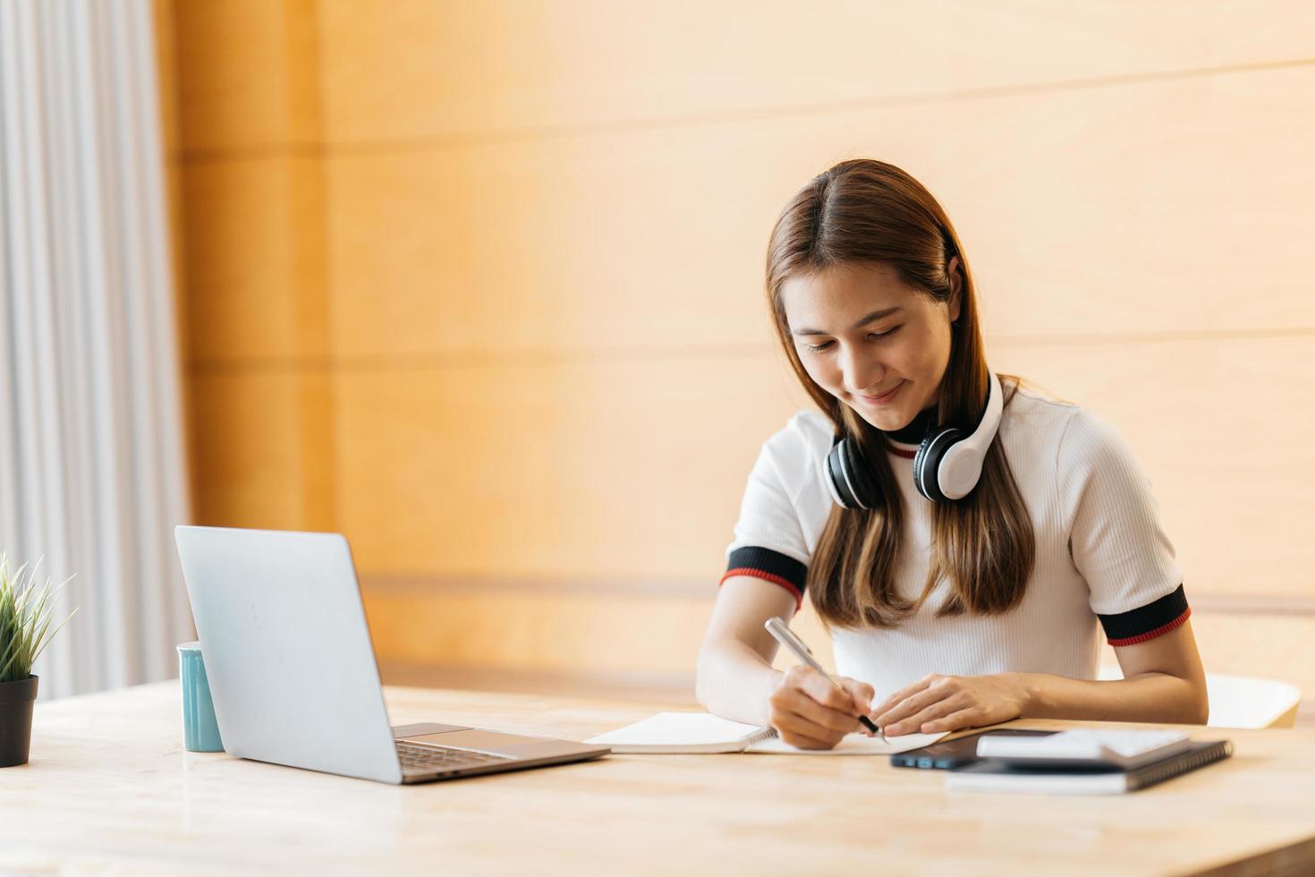 Happy asian businesswoman wearing headset make notes talk by conference call doing video chat on laptop in office, smiling chinese support service agent write information consult customer photo