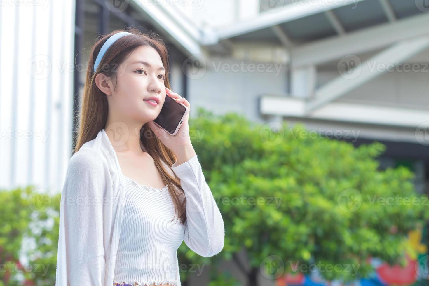 Young Asian business professional in white dress is calling phone seriously with office staff at outdoor building and bushes as the background. photo