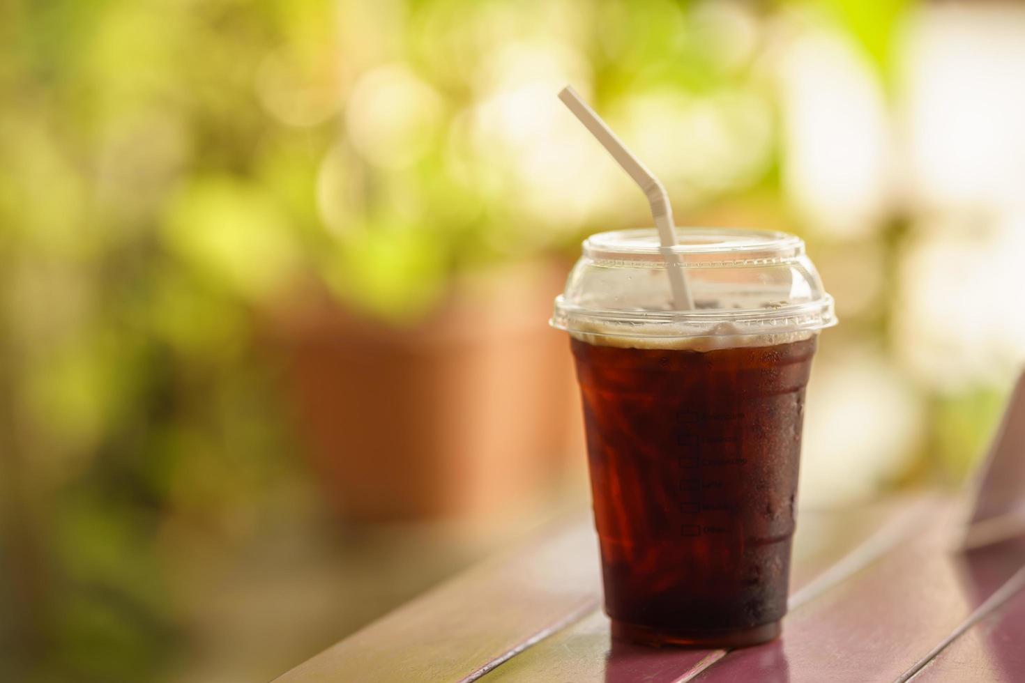 Closeup of takeaway plastic cup of iced black coffee Americano on wooden table with green nature background. photo