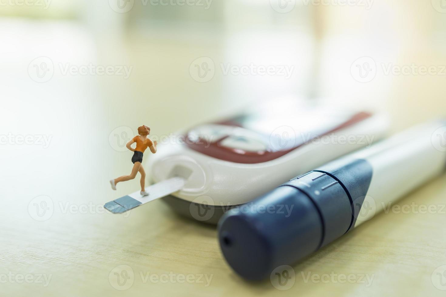 Medicina, diabetes, glucemia, atención de la salud y concepto de personas: cierre de una figura en miniatura de una corredora que se ejecuta en una tira de prueba de azúcar en la sangre y se conecta al medidor de glucosa en una mesa de madera. foto