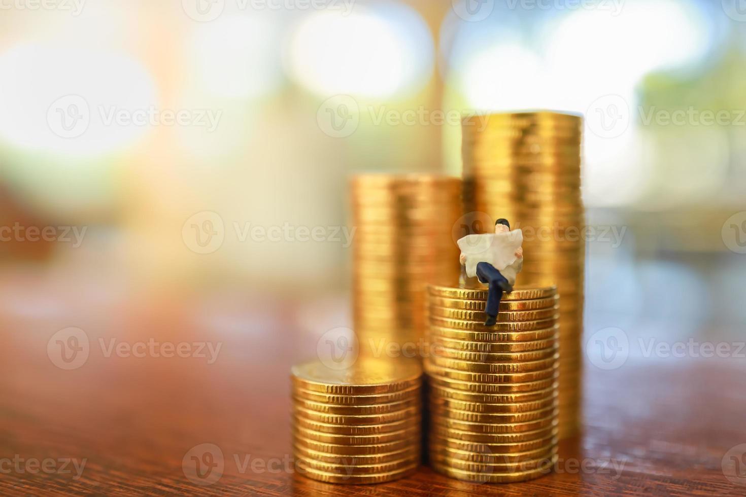 concepto de dinero, negocios, ahorro y planificación. primer plano de la figura en miniatura de un hombre de negocios sentado y leyendo un periódico en una pila de monedas de oro en una mesa de madera con espacio para copiar. foto