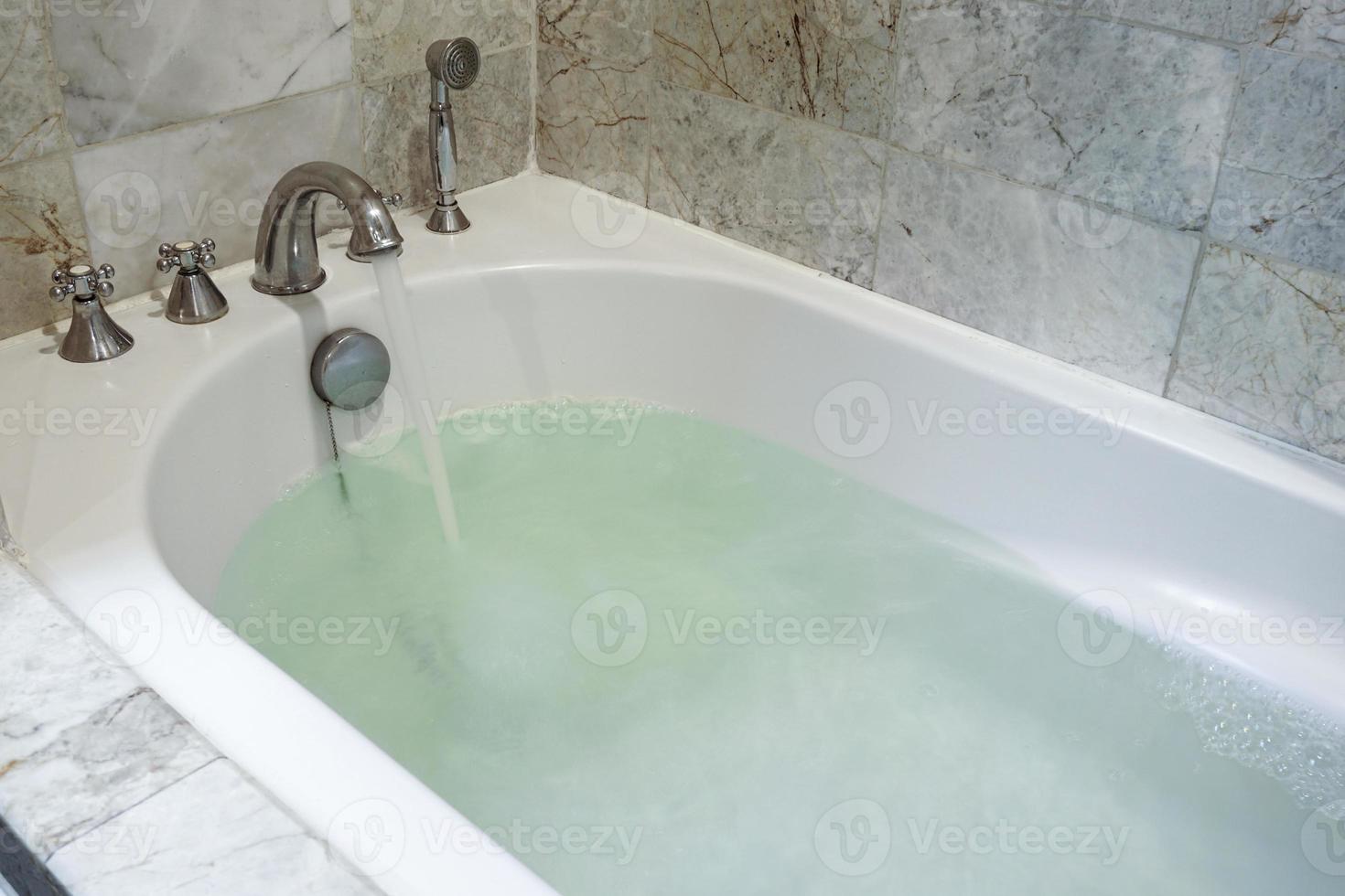 Stainless Faucet with a white bathtub that long large container filled with water in the bathroom decor with marble tile. photo