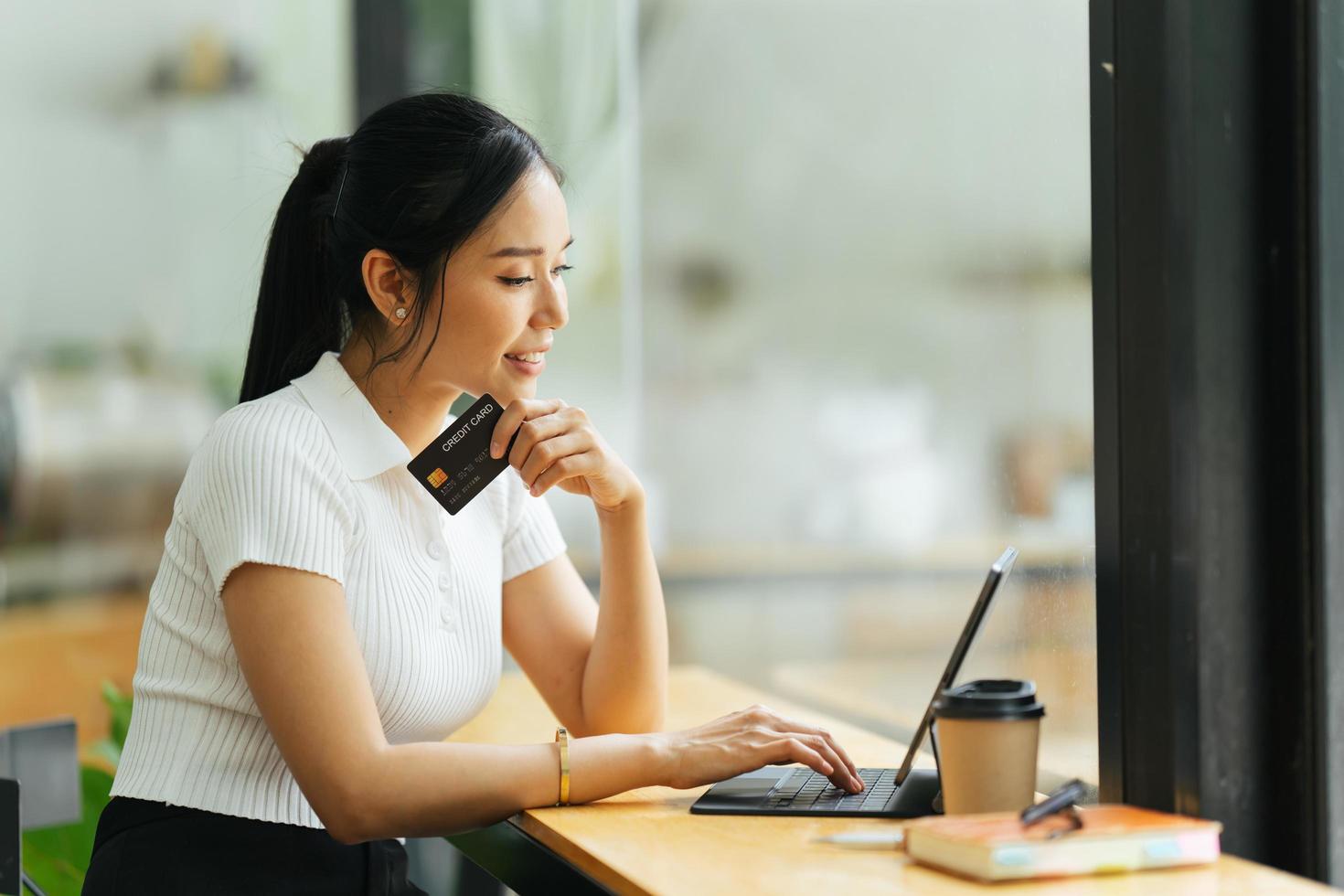 smiling woman paying online, using laptop, holding plastic credit card, sitting on coffee shop, Asian female shopping, making secure internet payment, browsing banking service. photo