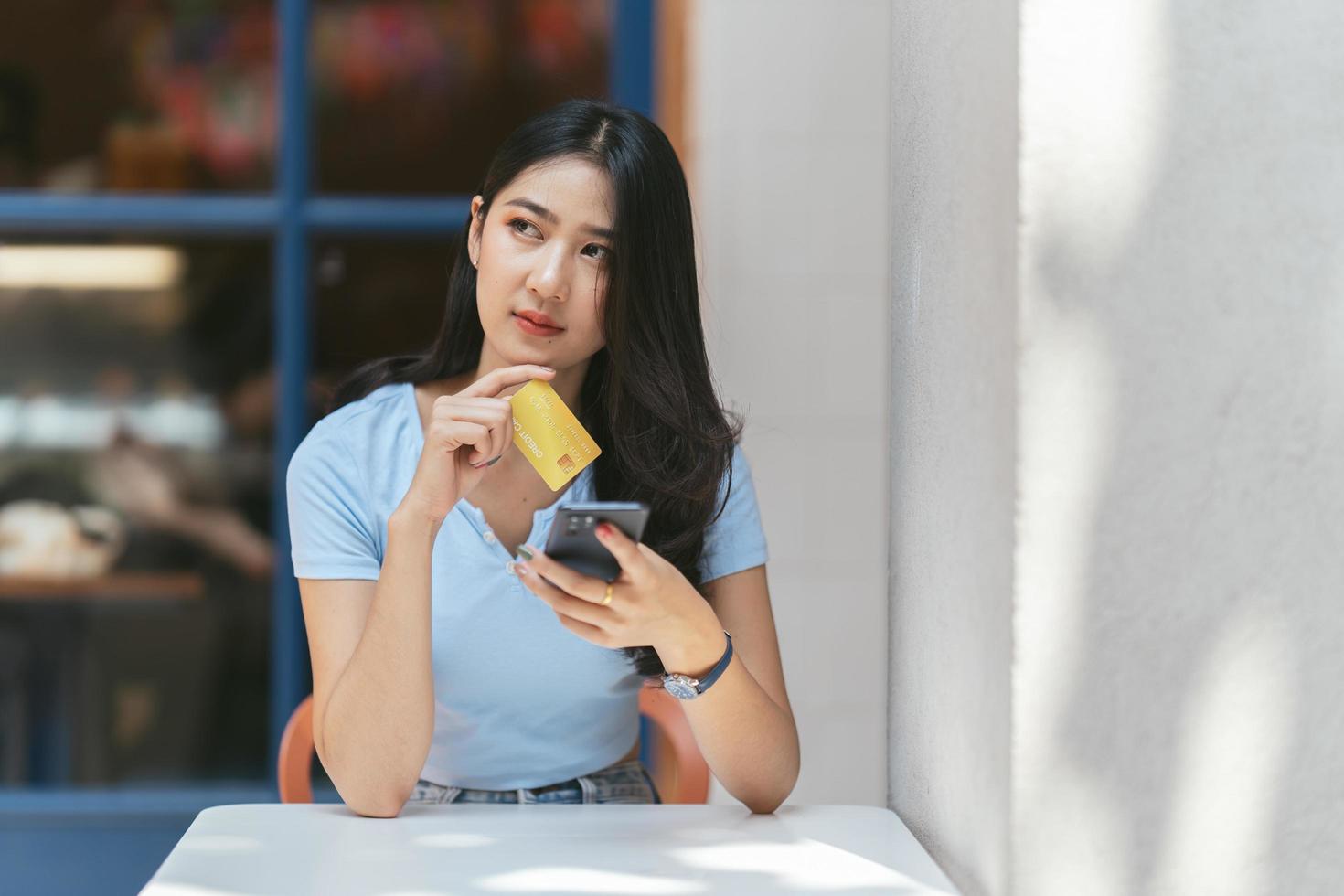 concepto de banca en línea. retrato de una joven asiática feliz con teléfono inteligente y tarjeta de crédito sentada en un café, mujeres asiáticas sonrientes disfrutando de hacer pagos desde casa. foto