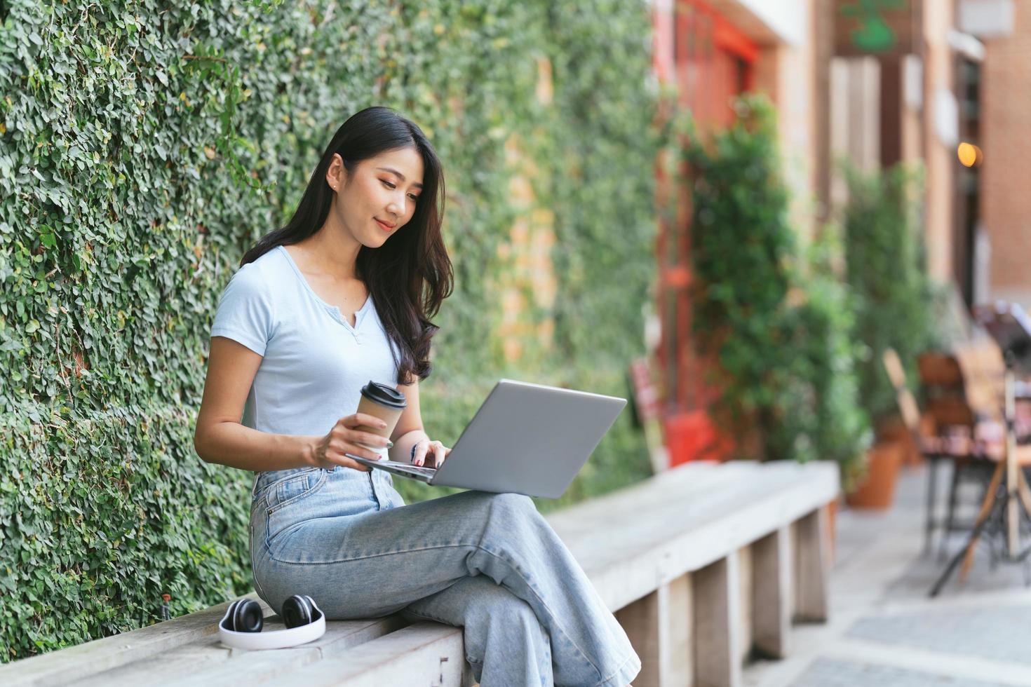 Portrait of beautiful Asian woman sitting outdoors at coffee shop restaurant during summer, using smart wireless technology computer laptop and smartphone, relaxing coffee break at cafe restaurant. photo