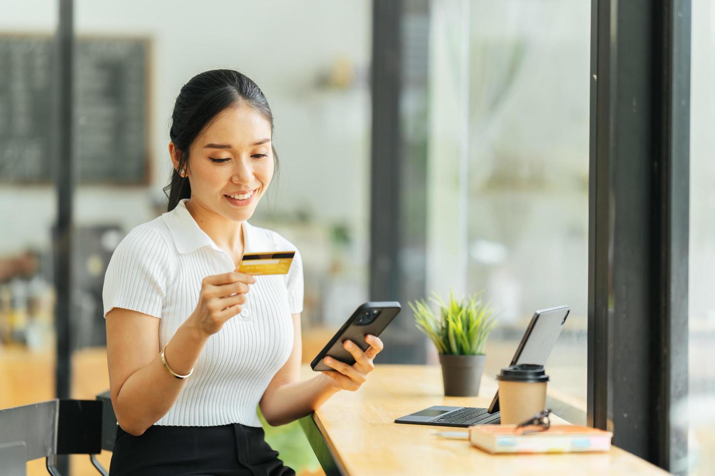 smiling woman paying online, using laptop, holding plastic credit card, sitting on coffee shop, Asian female shopping, making secure internet payment, browsing banking service. photo