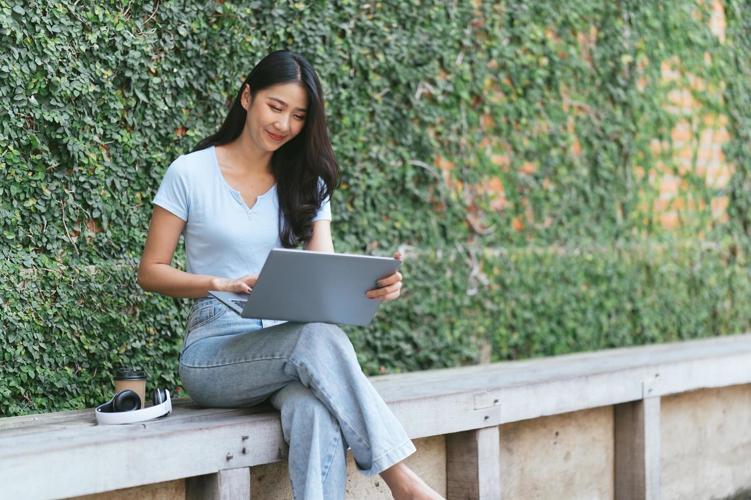 Portrait of beautiful Asian woman sitting outdoors at coffee shop restaurant during summer, using smart wireless technology computer laptop and smartphone, relaxing coffee break at cafe restaurant. photo