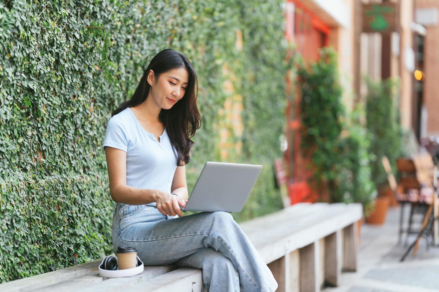 Portrait of beautiful Asian woman sitting outdoors at coffee shop restaurant during summer, using smart wireless technology computer laptop and smartphone, relaxing coffee break at cafe restaurant. photo