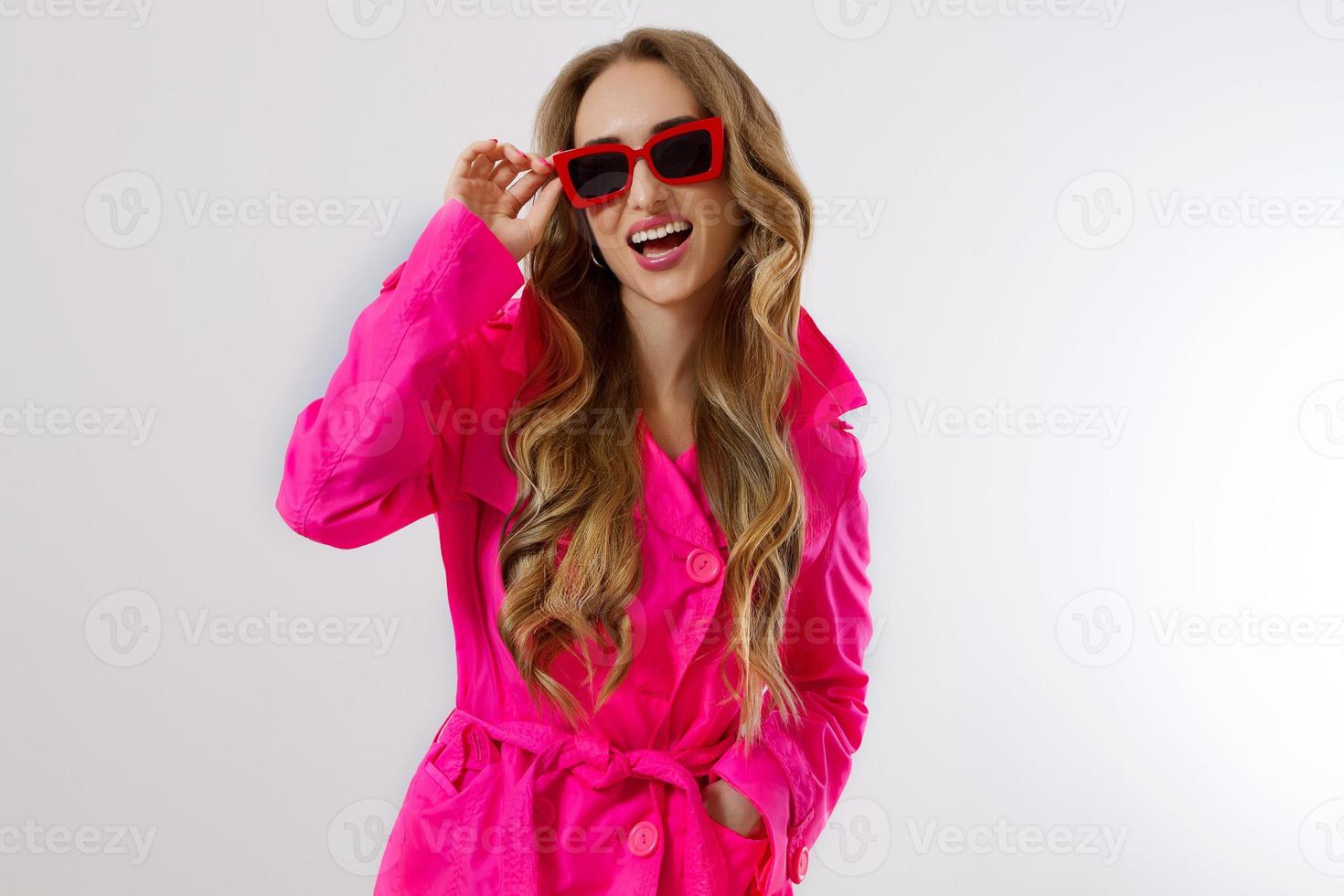 Cierra a una chica feliz con gafas de sol rojas y un abrigo rosa aislado de fondo blanco. mujer joven emocionada, conmocionada y sorprendida con ropa de moda. concepto de moda y compras. pelo claro largo y ondulado. foto
