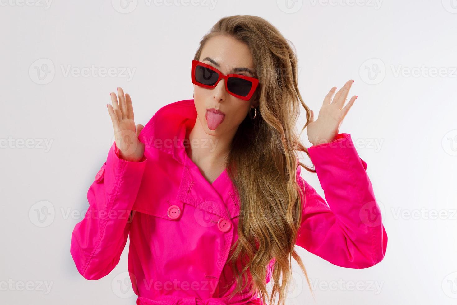 cara de chica loca y graciosa con la lengua fuera. mujer feliz con cara divertida aislada sobre fondo blanco. bella dama con abrigo rosa y gafas de sol rojas. traje de moda y de moda. modelo de glamour foto