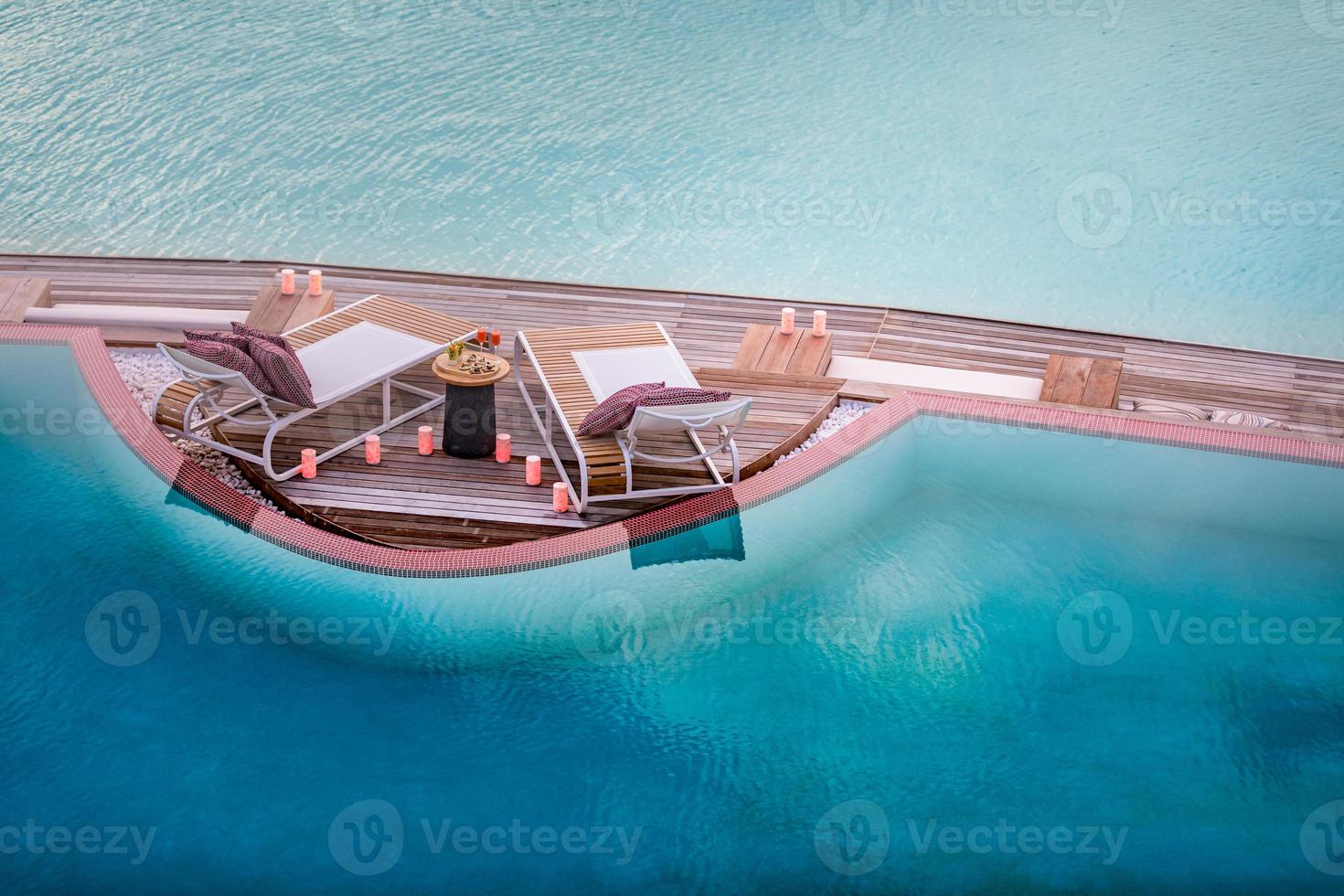 Summer love and romance table set-up for a romantic dinner meal with infinity pool reflection chairs under sunset sky and sea in the background. Luxury destination dining, romantic dinner for couples photo