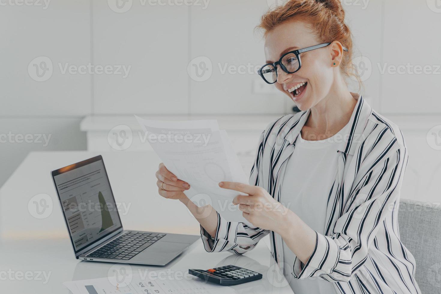 mujer pelirroja encantada leyendo documento financiero y sonriendo de noticias agradables foto