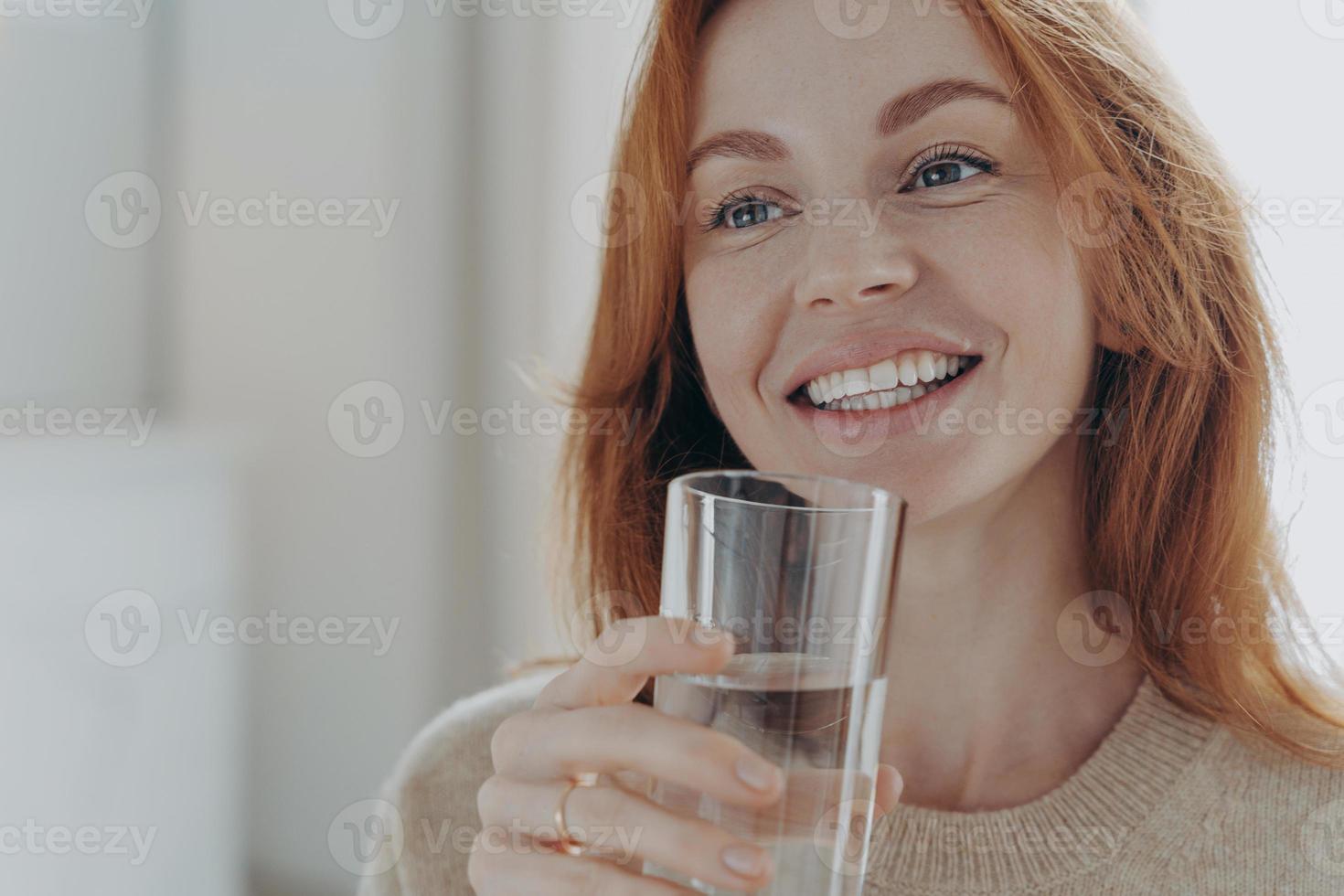 retrato de una feliz y saludable mujer pelirroja sosteniendo un vaso transparente de agua mineral pura foto