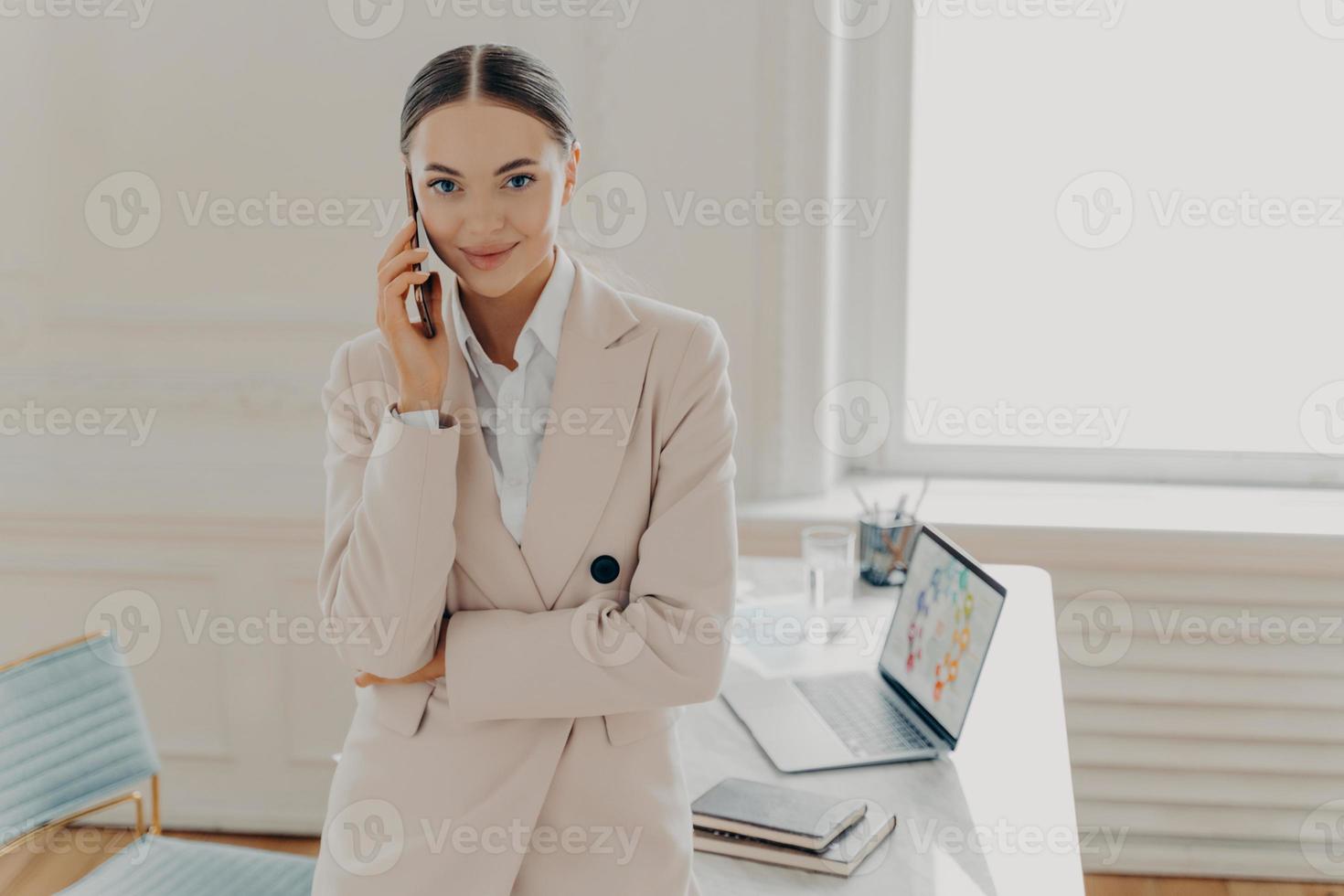 Positive young female entrepreneur talking on phone and smiling at camera photo