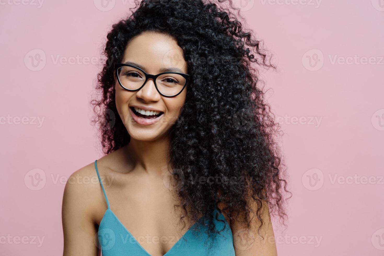 Close up shot of happy carefree African American woman laughs from joy, has healthy dark skin, curly hair, isolated over pastel rosy background, hears compliment. People, emotions, happiness concept photo