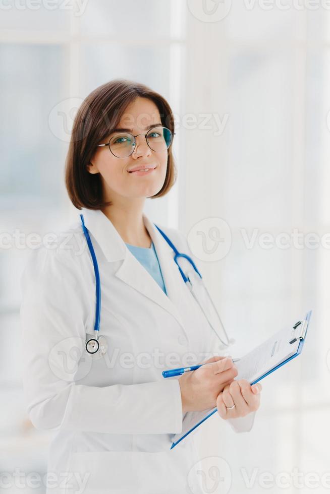 Qualified cardiac expert with stethoscope prepares for health seminar, gives consultancy about first aid, writes down information in clipboard, wears white gown, ready to give medic care help photo