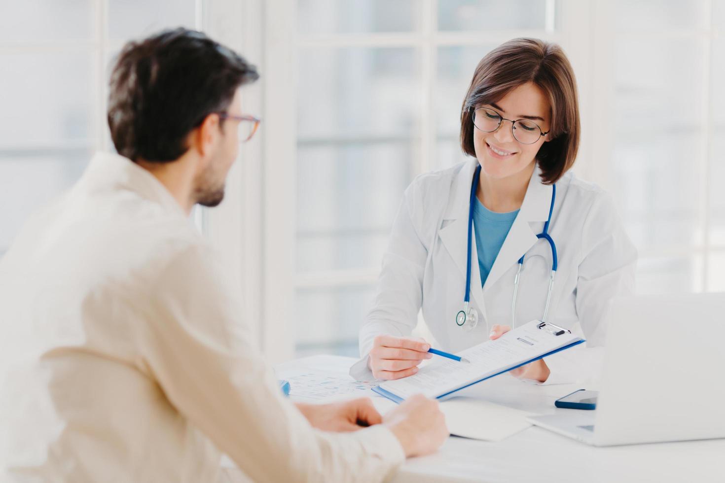 Medical appointment. Female doctor gives professional medical help to male patient, explains written information on paper in clipboard, gives support and good service, pose at hospital near desktop. photo