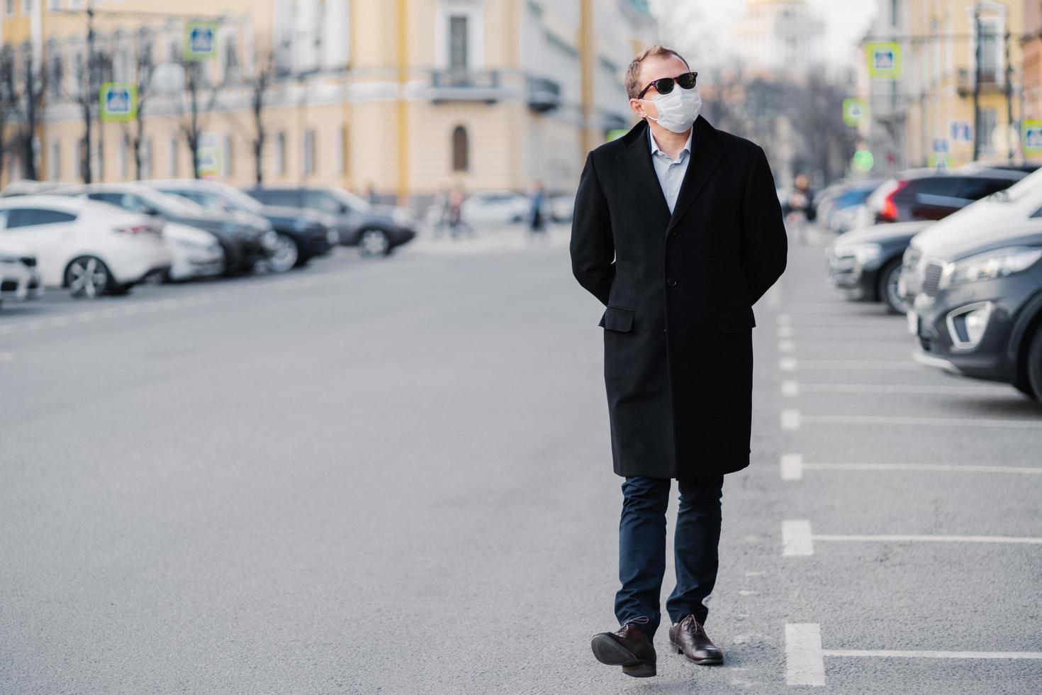 Full length shot of serious man walks at street, dressed in elegant outfit, wears medical mask to prevent coronavirus or another type of virus, stays safe during quarantine time. Pandemic situation photo